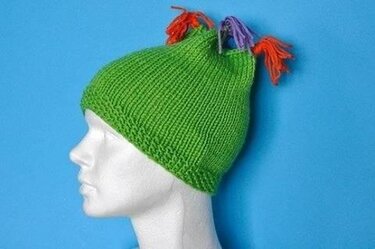 Schaufensterbüste mit grüner gestrickter Damenmütze und drei bunten Quasten vor blauem Hintergrund