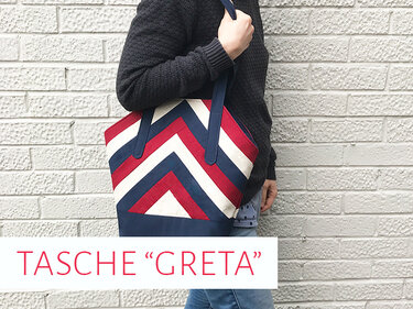 Person trägt Stofftasche mit Zickzack-Muster in Rot, Weiß und Dunkelblau vor einer hellen Ziegelwand.