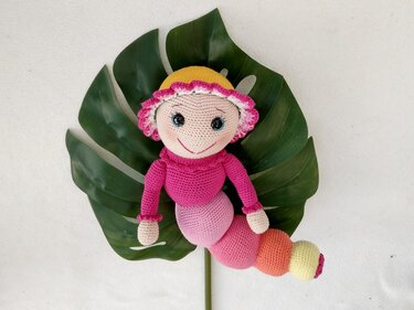Gehäkelte Raupenpuppe in Pink und Rosa mit gelben Akzenten, liegend auf einem großen grünen Monstera-Blatt