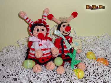 Zwei fröhliche Amigurumi-Käfer in Rot und Schwarz mit Schleifen und gehäkelter Blume auf weißer Häkeldecke