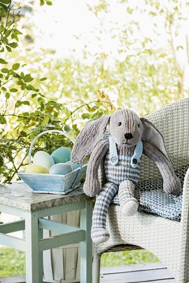 Gestrickter Amigurumi-Hase in gestreifter Latzhose auf Korbstuhl neben Korb mit pastellfarbenen Ostereiern.