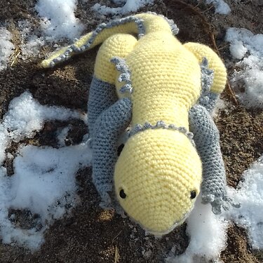 Gelbe gehäkelte Amigurumi-Eidechse mit grauen Beinen und Rückenkamm auf leicht verschneiter Erde.
