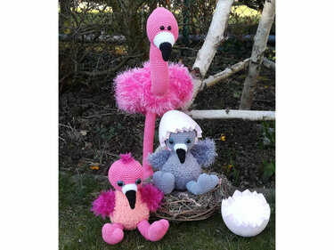 Three crocheted flamingo amigurumi toys: tall pink adult, small pink chick, and grey chick in eggshell sitting in a nest on grass by birch branches