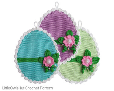 Three pastel egg-shaped crochet potholders in blue, purple and green with white scalloped edges and pink flower appliqués with bead centers
