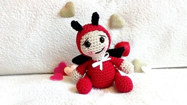 Gehäkelte Amigurumi-Glückskäfer-Puppe rot/beige mit schwarzen Fühlern und Flügeln auf weißem Stoff, umgeben von kleinen Herzen.