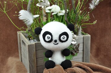 Gehäkeltes schwarz-weißes Panda-Amigurumi sitzt vor einer Holzkiste mit Blumen und Federn.