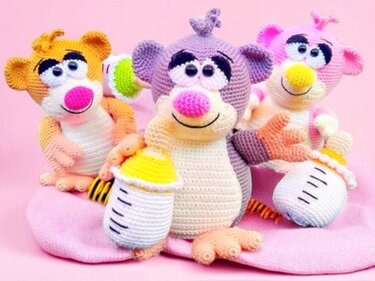 Drei gehäkelte Amigurumi-Tiere (orange, lila, rosa) mit Babyflaschen auf rosa Decke.