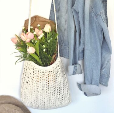 Cremefarbener Häkel‑Shopper mit Ajourmuster, gefüllt mit Blumen und neben einer Jeansjacke hängend