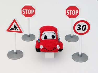 Rotes gehäkeltes Amigurumi-Auto umgeben von vier Spielzeug-Verkehrsschildern auf weißem Hintergrund.