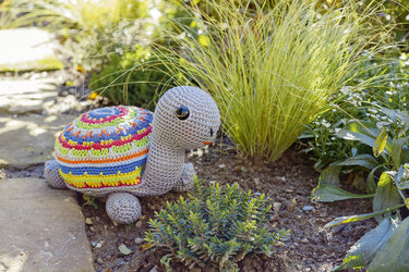 Gehäkelte Amigurumi-Schildkröte mit grauem Körper und buntem gestreiftem Panzer zwischen Gartenpflanzen.