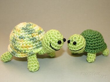Zwei Amigurumi-Schildkröten, eine größere mit meliertem Panzer und eine kleinere in Dunkelgrün, beide mit aufgestickten Mündern und schwarzen Augen.