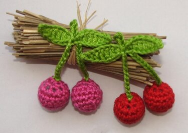 Vier gehäkelte Kirschen (zwei pink, zwei rot) mit grünen Blättern auf einem Bündel Stroh.