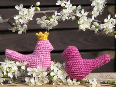Zwei rosa Amigurumi-Häkelvögel, einer mit gelber Krone, zwischen weißen Blüten auf Holzhintergrund