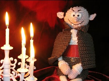 Gehäkelter Amigurumi‑Vampir mit Umhang und roter Schärpe neben einem weißen Kerzenleuchter mit brennenden Kerzen.