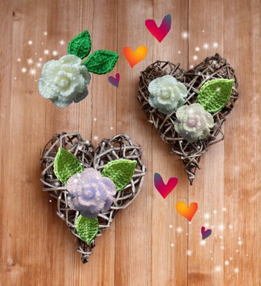 Drei gehäkelte Blumen mit grünen Blättern, zwei befestigt auf Weidenherzen, vor hellem Holzhintergrund mit funkelnden Akzenten.