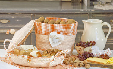Ländliche Tischdekoration mit zwei genähten Körben, gefüllt mit Brot und Kartoffeln, umgeben von Trauben, Käse, Nüssen und einer Keramikkanne.