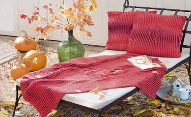 Rote gestrickte Decke und zwei Kissen mit Blattmotiv auf einer Liege, umgeben von Kürbissen und grüner Glasvase