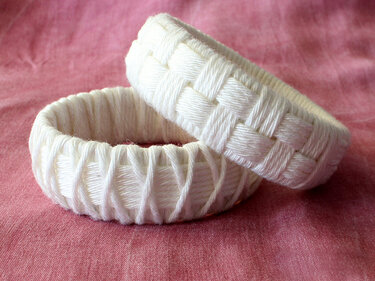 Two white yarn-wrapped bangles showing basket-weave and diagonal wrap styles on pink fabric