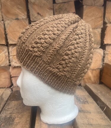 Beige gestrickte Mütze mit Zopfmuster auf Schaumkopf vor Holzstapel