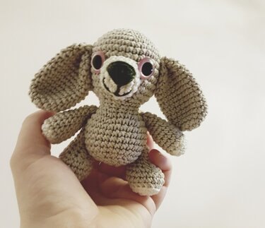 Kleiner beiger gehäkelter Amigurumi-Hund mit großen Schlappohren und Wackelaugen in einer Hand.