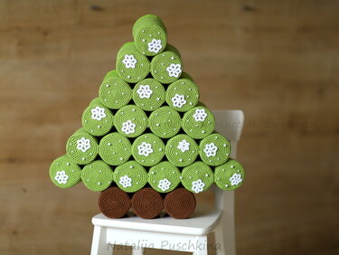 Gehäkelter Tannenbaum aus grünen Rollen mit weißen Blumenapplikationen und brauner Basis auf einem weißen Stuhl vor Holzwand