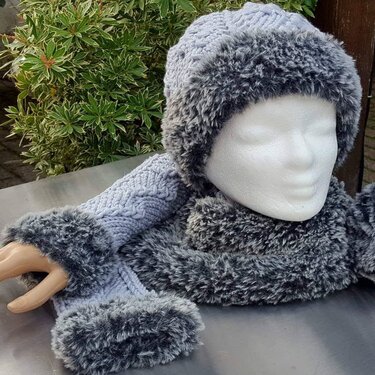 Strickset aus Mütze, Schal und Armstulpen in blaugrauem Zopfmuster mit dunklem Kunstfellbesatz, präsentiert auf einer Schaufensterpuppe.