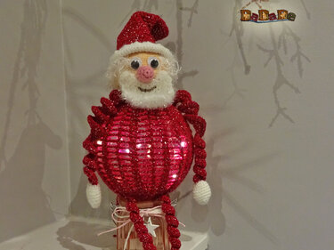 Gehäkelter Weihnachtsmann mit rotem Glitzerkörper und weißem Bart, sitzend auf einem Glasgefäß.