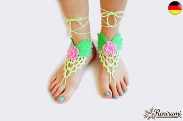Zwei Füße tragen hellgrüne gehäkelte Barfußsandalen mit rosa Blüte und grünen Blättern, am Knöchel geschnürt.