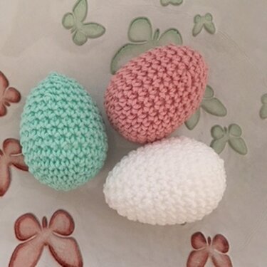 Drei gehäkelte Ostereier in Mintgrün, Rosa und Weiß auf einem Stoff mit Schmetterlingsmotiv.