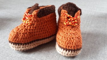 Orangene gehäkelte Babyschuhe mit braunem Rand und gelb-roter Schnürung auf grauem Stoff