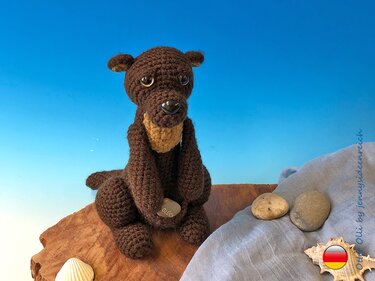 Brauner Amigurumi-Otter mit hellem Brustteil hält Stein „Glück“, sitzt auf Holzscheibe neben Tuch, Steinen und Muscheln