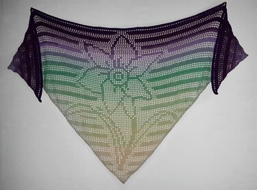 Häkel-Dreieckstuch im Filetmuster mit großem Blütenmotiv und Lila‑Grün‑Beige Farbverlauf, an der Wand drapiert