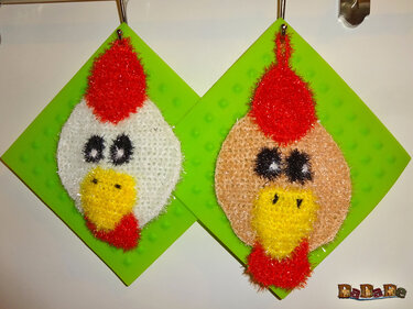 Zwei gehäkelte Hühnerkopf-Spüllappen, weiß und hellbraun, mit roten Kämmen und gelben Schnäbeln, an Haken aufgehängt