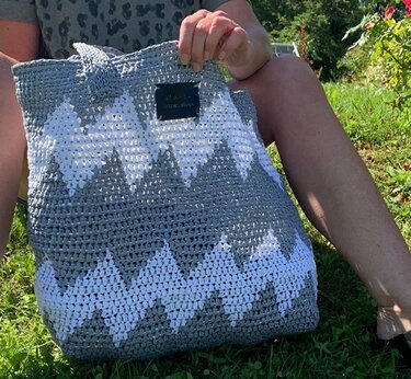 Gehäkelter grau-weißer Shopper mit Zickzackmuster, gehalten von einer Person auf einer Wiese