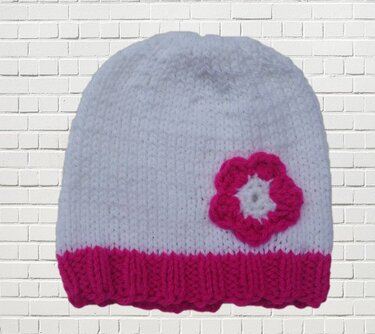 Weiße gestrickte Babymütze mit pinkem Rippenbündchen und pinker Blumenapplikation vor weißer Ziegelwand