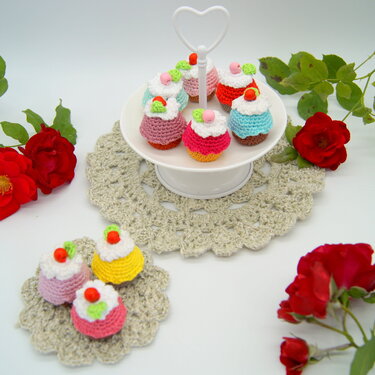 Weiße Etagere mit bunten gehäkelten Mini-Törtchen; zusätzliche Törtchen auf einem gehäkelten Deckchen und rote Rosen als Dekoration.