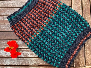 Gestrickter Loop-Schal in Dunkelblau, Petrol und Orange mit Mosaikmuster auf Holzuntergrund neben roten Blüten