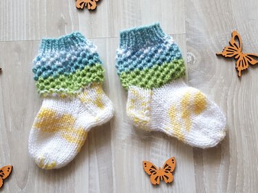 Nahaufnahme zweier gestrickter Babysocken mit strukturiertem blau-grünem Schaft und weißem Fuß mit gelben Einsprengseln auf Holzuntergrund