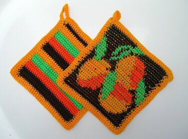 Zwei gehäkelte quadratische Topflappen mit gelb-orangenem Rand, ein Topflappen mit gestreiftem Muster, einer mit Früchtemotiv und Aufhängeschlaufen