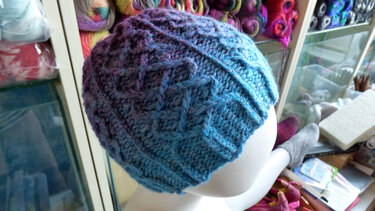 Gestrickte Beanie in Blau-Lila mit Zopfmuster auf Mannequinkopf vor Regalen mit Wolle
