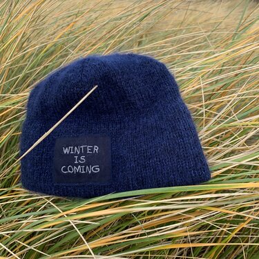 Dunkelblaue gestrickte Beanie mit 'WINTER IS COMING'-Patch und einem dünnen Holzstab im Dünengras.