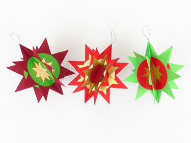 Drei 3D-Papiersterne in Rot, Grün und Gold mit goldenen Aufhängeschlaufen auf hellem Hintergrund.