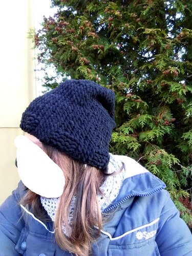 Person trägt grob gestrickte dunkle Mütze und hellgrauen Loop‑Schal vor Nadelbaum
