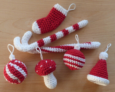Gehäkelte rote und weiße Weihnachtsanhänger (Zuckerstangen, Mütze, Stiefel, Kugeln, Pilz) auf einer Holzoberfläche.