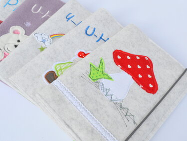 Mehrere Filzhüllen mit applizierten Motiven (Pilz, Bär, Regenbogen) als Geschenkideen zur Geburt