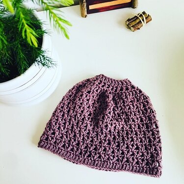 Lila gehäkelte Mütze mit Öffnung für Pferdeschwanz auf weißer Fläche neben Zimmerpflanze.