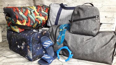Mehrere Reisetaschen und Rucksäcke in unterschiedlichen Mustern und Farben, dazu blaue Badesandalen, Schwimmbrille und ein Spielzeugring.