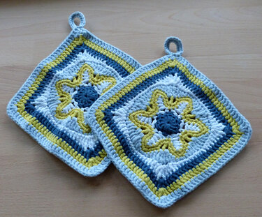 Zwei quadratische, handgehäkelte Topflappen in Blau, Gelb und Weiß mit Sternmuster und Aufhängeschlaufen auf Holz.
