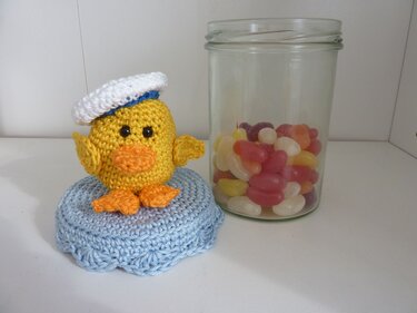 Gelb gehäkeltes Küken mit weißer Matrosenmütze und orangefarbenem Schnabel auf hellblauer gehäkelter Unterlage neben einem Glas mit bunten Geleebohnen.