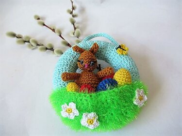 Gehäkelter Osterkranz: hellblauer Ring mit grünem, flauschigem Bereich, mittig ein brauner Hase, davor bunte Ostereier und weiße Häkelblüten, kleiner gelber Schmetterling